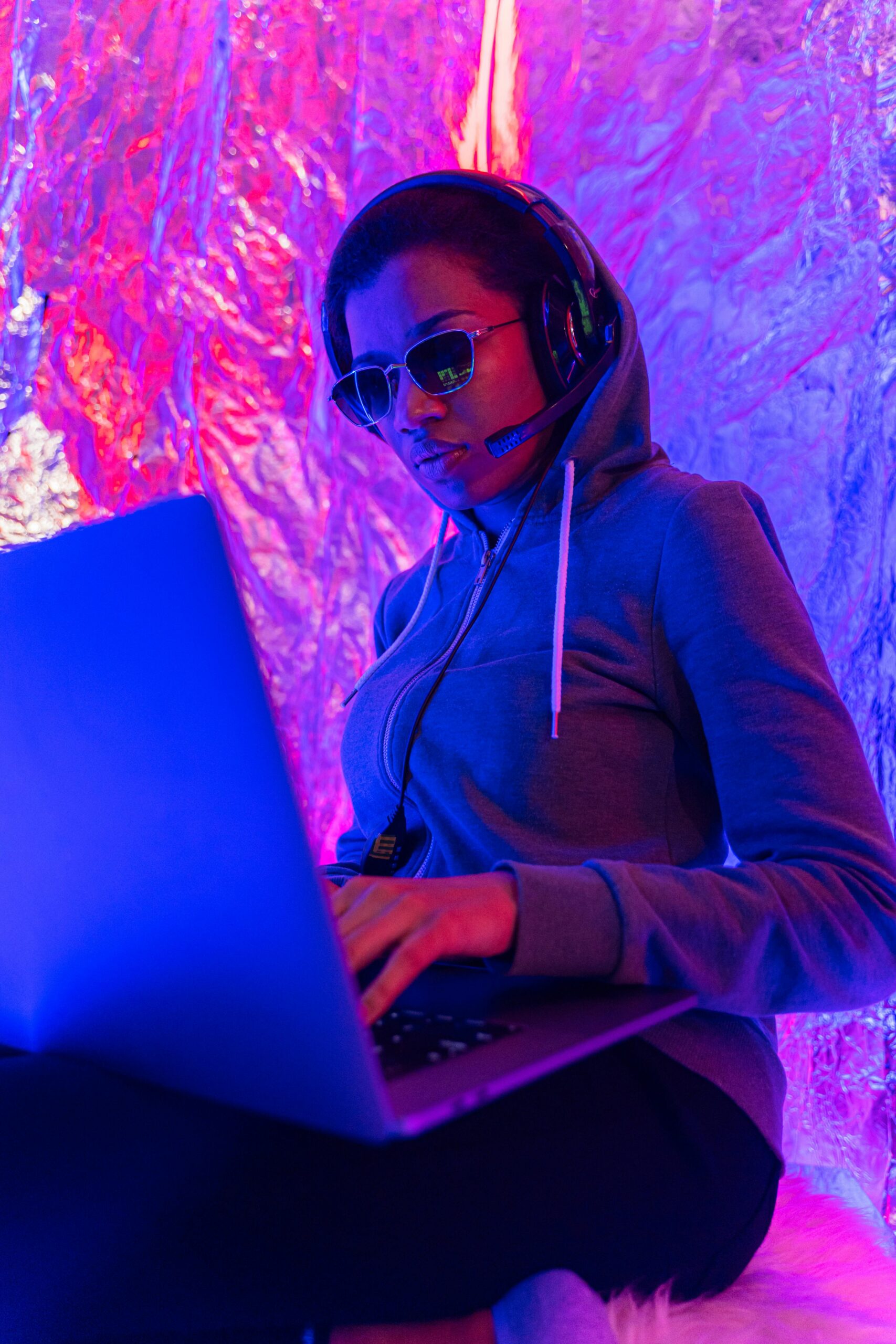 Woman in hoodie and headphones engaging with a laptop in a vibrant, futuristic setting.