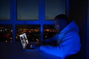 Man working on a laptop at night by a city-lit window, focusing on technology.
